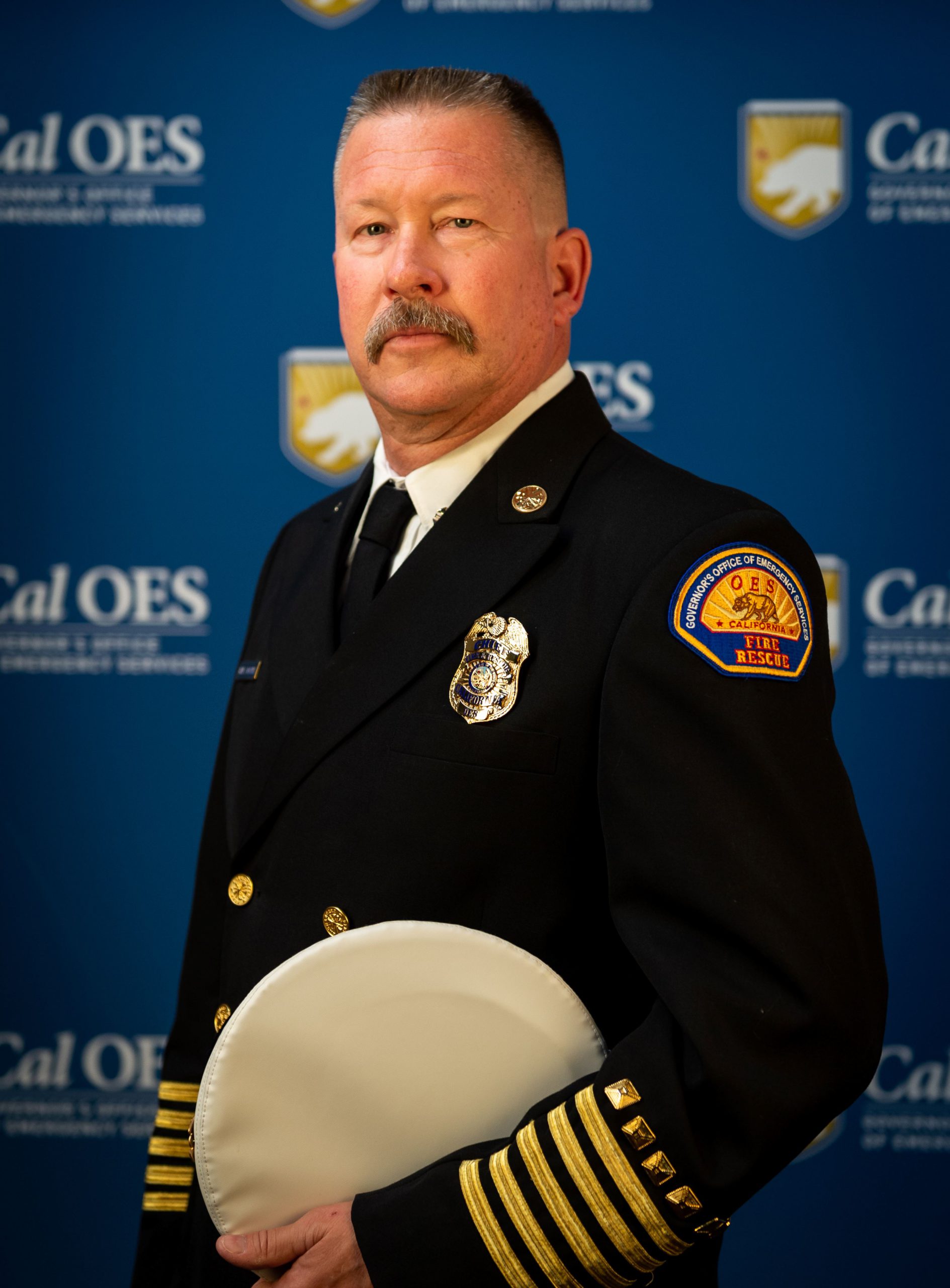 Cal OES Fire and rescue Chief Brian Marshall holding his hat. Posed in front of Cal OES background in full uniform.