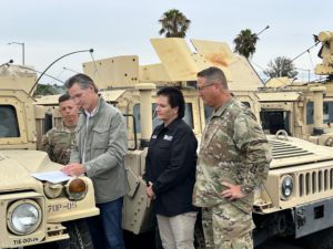 Governor Newsom signs a document on the hood of a military vehicle with military personnel and cal O E S employee standing beside watching.