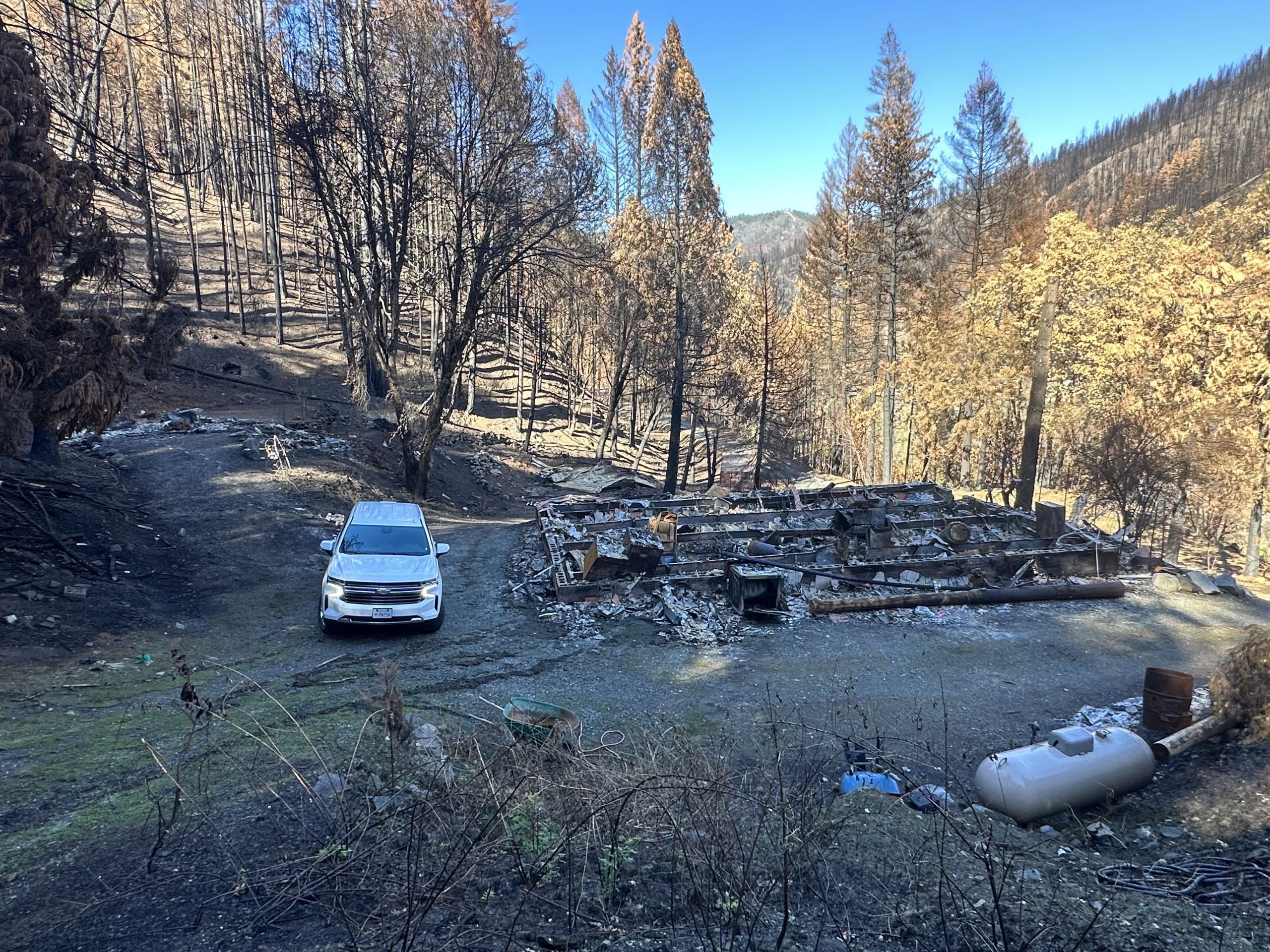 A burned structure base surrounded by tall pine trees. A Cal O E S vehicle is next to it.