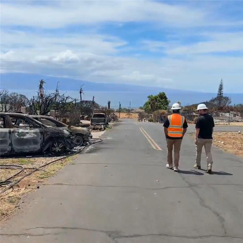 Two Cal OES employees walk along a road on Maui. The impacts of the Lahaina wildfire, including burned vehicles, are visible around them.