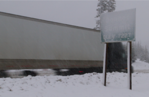 A big rig with a dark cab and white trailer drives through snow on I-80 past the Donner Summit sign, which is covered in snow.