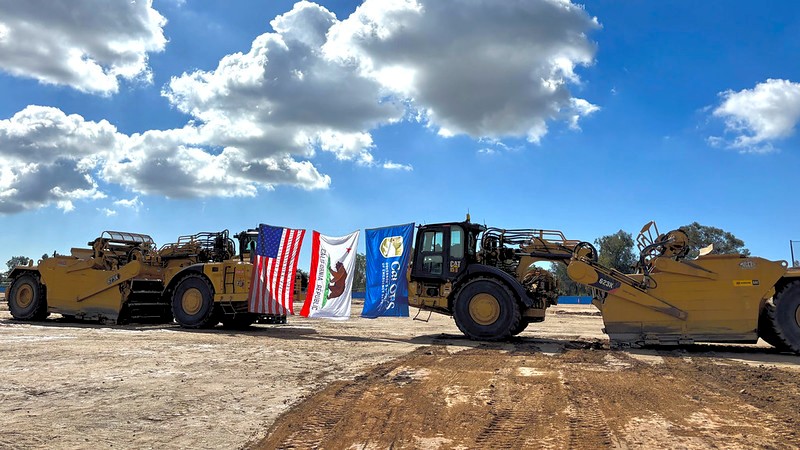 A U.S. flag, state of California flag and Cal OES flag suspended between two large construction vehicles.